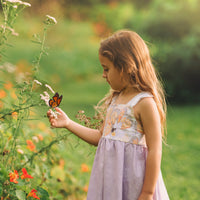 A young girl wearing a sleeveless dress with a Monarch butterfly print and lace, standing in a garden.