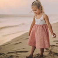 A young girl standing on a beach wearing a white sleeveless dress with a sailboats print and a twirly skirt.