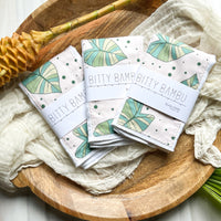 Three folded baby burp cloths with taro/kalo print design on a wooden surface with a white bamboo mat and a green leaf, with the brand name 'bitty bambu' visible on the cloths.