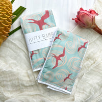 A single baby burp cloth with an Iwa bird design, laid flat on a white textured surface, next to a flower and a decorative palm leaf.