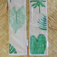 Two beige baby burp cloths with green watercolor leaf designs laid out on a wicker surface.
