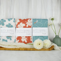 Three folded burp cloths with hibiscus print design, displayed on a wooden tray with a white fabric backdrop and a vase with flowers to the side.