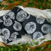 A set of black and white opihi shell patterned organic knit shorts for babies, displayed on a wooden surface with green leaves and a white fabric in the background.