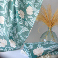 A swaddle blanket with a floral pattern in shades of green, draped over a white surface, alongside a decorative vase with dried flowers.