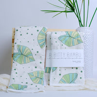 Two burp cloths with taro leaf pattern on a white surface with a plant in the background