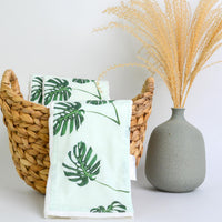 Towels with leaf pattern in a basket next to a vase on a white background
