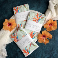 Folded burp cloths with a tropical bird of paradise flower pattern, displayed alongside orange flowers on a textured dark background.