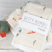 Packaged baby burp cloth with a floral pattern, displayed alongside a branding card with 'BITTY BAMBU' written on it, on a wooden surface.