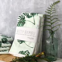 A folded white burp cloth with green monstera leaf patterns, displayed on a wooden surface with a decorative fern and a glass bottle in the background, suggestive of a baby gift theme.