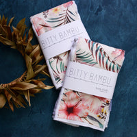 Folded baby burp cloths with a floral and tropical print design, placed on a dark textured surface next to a dried plant.