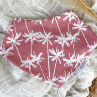 A pink bandana-style baby bib with white palm tree patterns laid out flat on a neutral fabric surface.