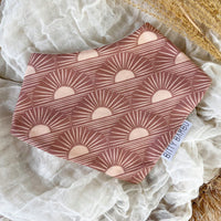 A brown bandana-style baby drool bib with a sun and geometric pattern, displayed on a flat surface with a beige backdrop.