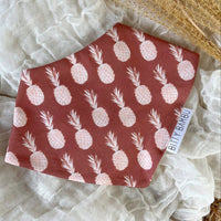 A brown bandana-style baby drool bib with pineapple print, displayed on a flat surface.