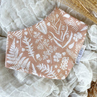 A beige baby drool bib with a bandana style, featuring a floral pattern with ferns, placed on a soft fabric background.