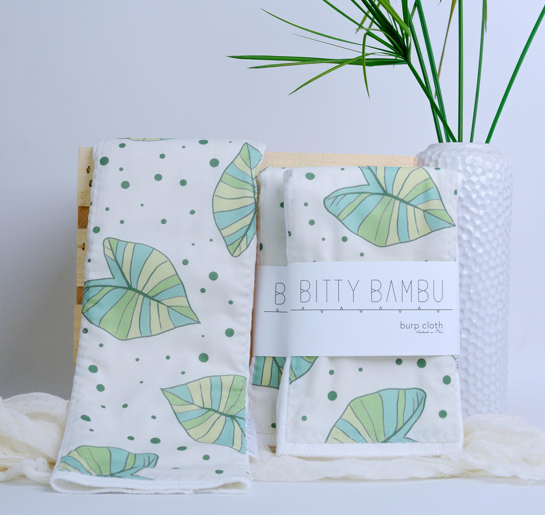 Two burp cloths with  taro leaf pattern on a white surface with a plant in the background